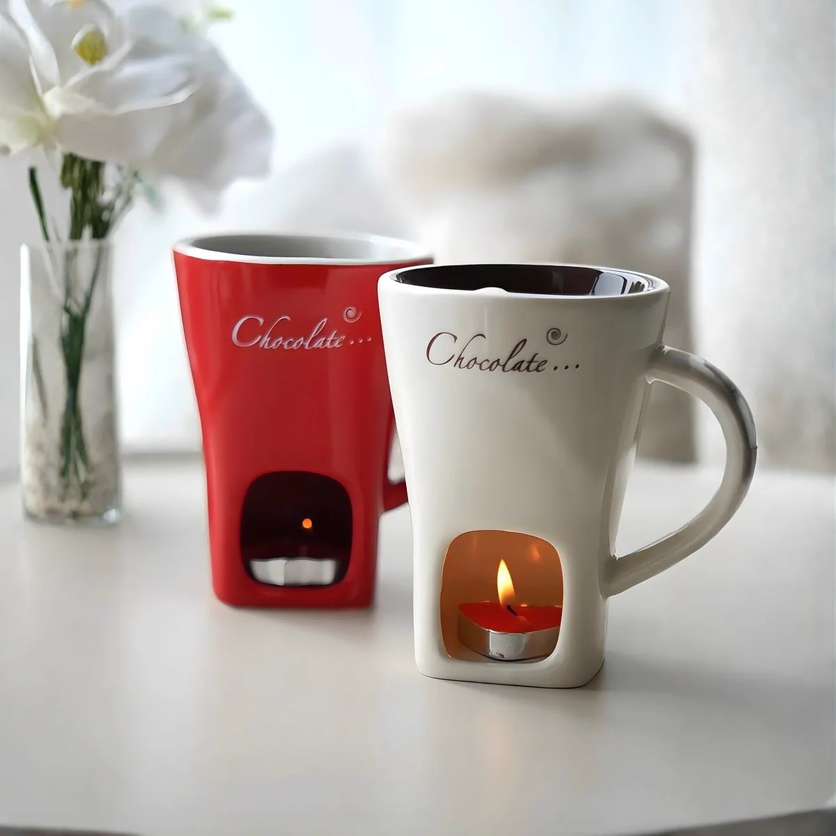 A white mug and a red mug side by side on a table, the tea light in the white one lit and glowing through the arch.