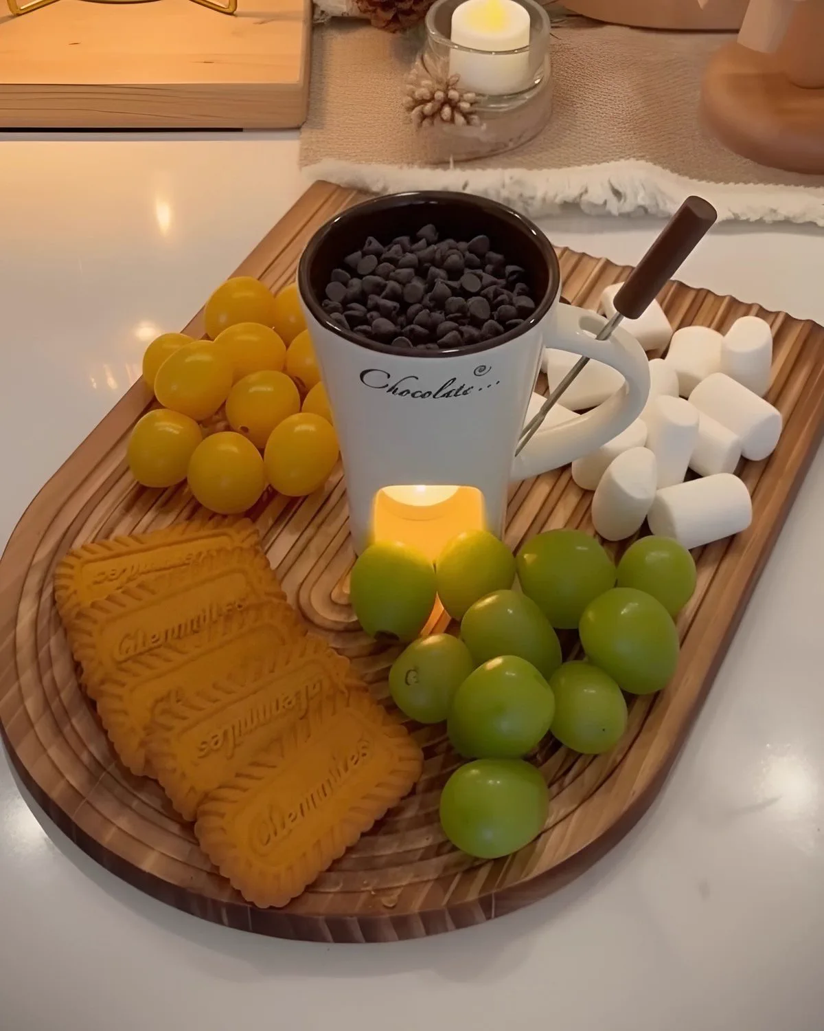 White fondue mug filled with chocolate chips on a wooden board with marshmallows, grapes and biscuits, a tea light glowing underneath.